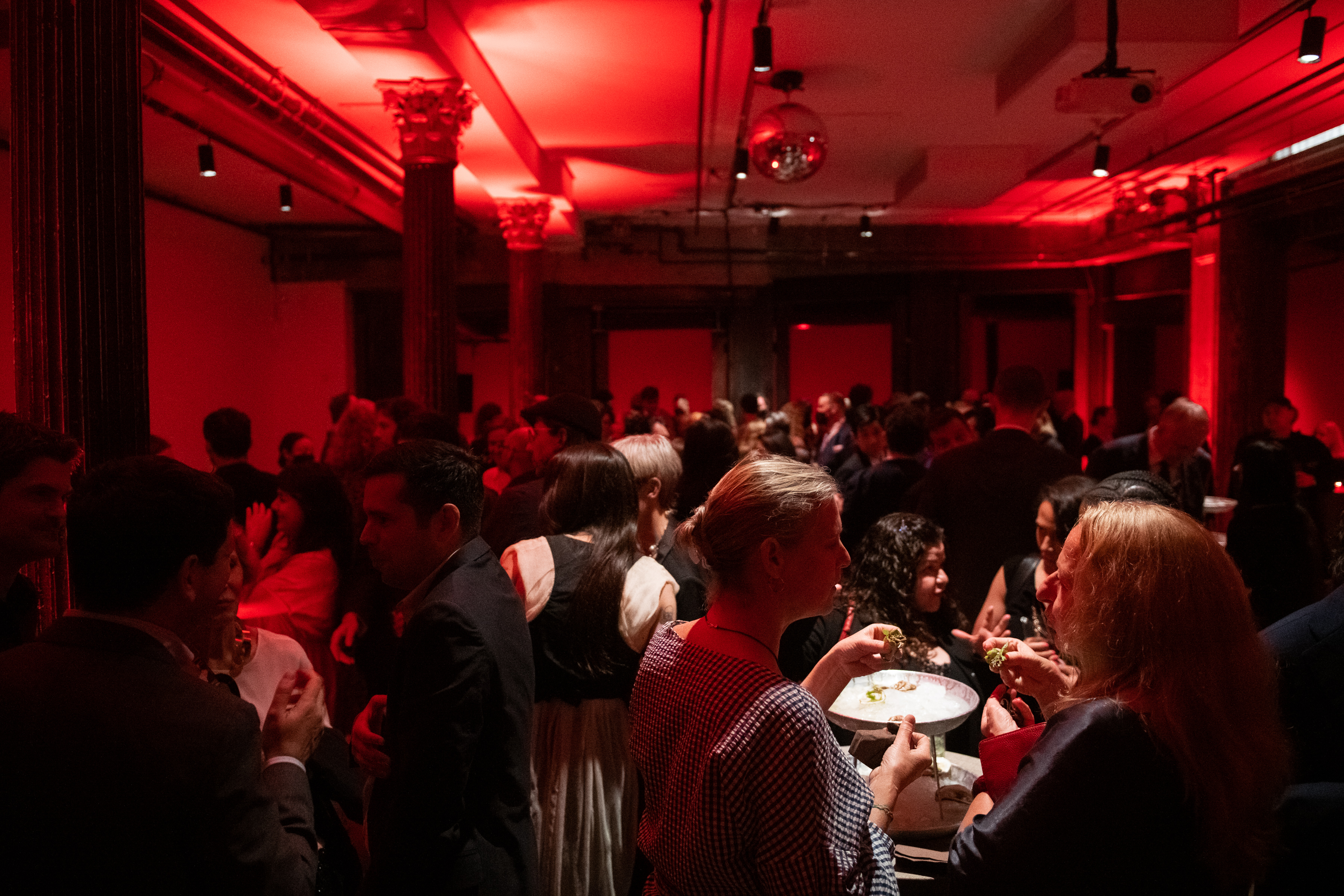 A color photo of several individuals engaged in festivities and banter within a large reception space. Light and dark red overhead lights shine onto the guests in the event.