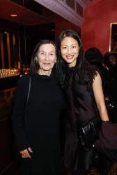 Two women stand close together and smile at the camera. Barbara Gladstone, who stands on the left, is wearing a black long-sleeved cowl-neck gown. Emily Wei Rales, who is on the right, wears a dark eggplant dress with fringe accents and trim.