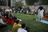 A group of people gathered outside in a semicircle around a performer. Some sit on benches, others on grass, and others stand. The performer is dressed in a long white robe and knee-length, high-heeled boots; their face is painted blue and they are speaking into a microphone.