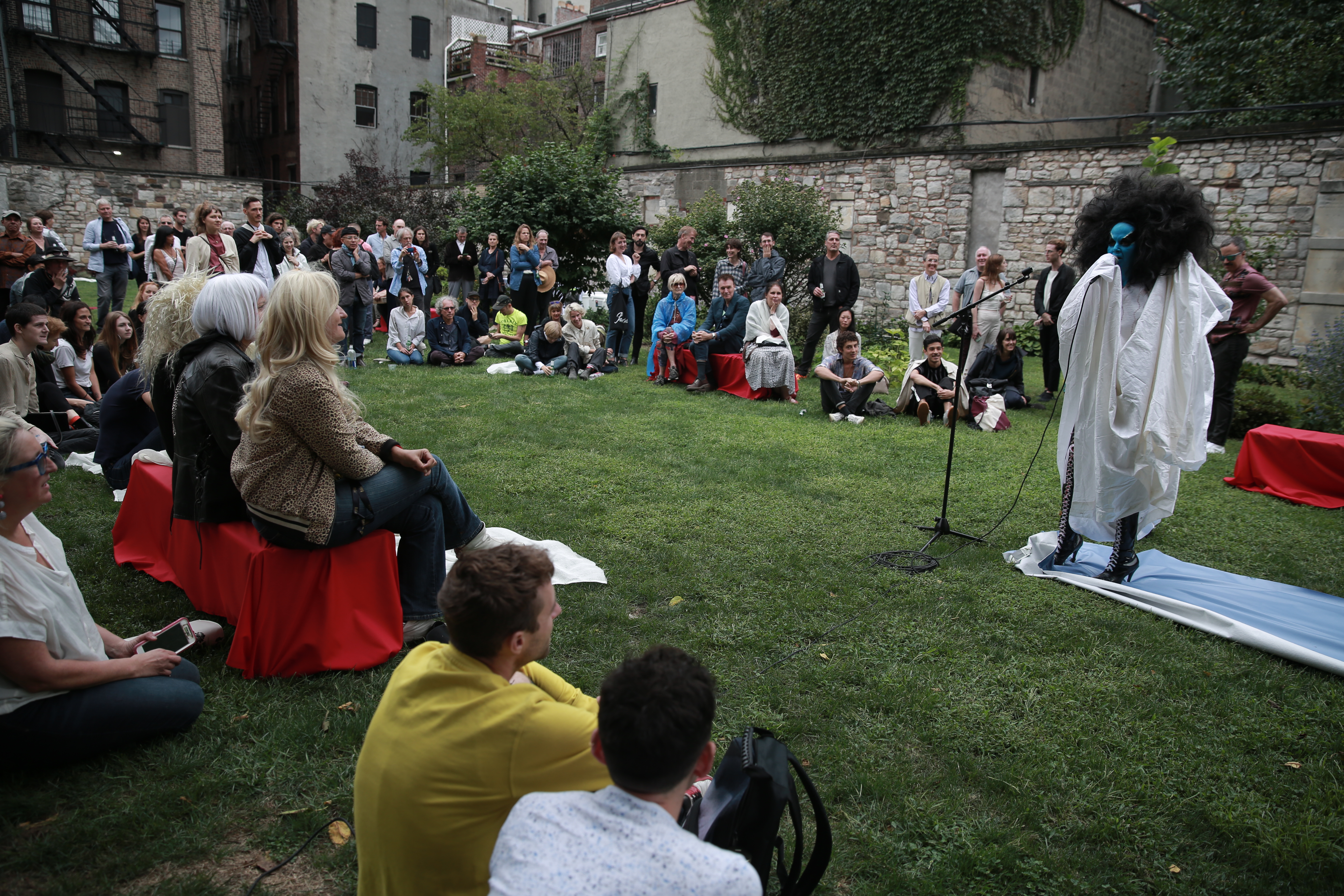 A group of people gathered outside in a semicircle around a performer. Some sit on benches, others on grass, and others stand. The performer is dressed in a long white robe and knee-length, high-heeled boots; their face is painted blue and they are speaking into a microphone.