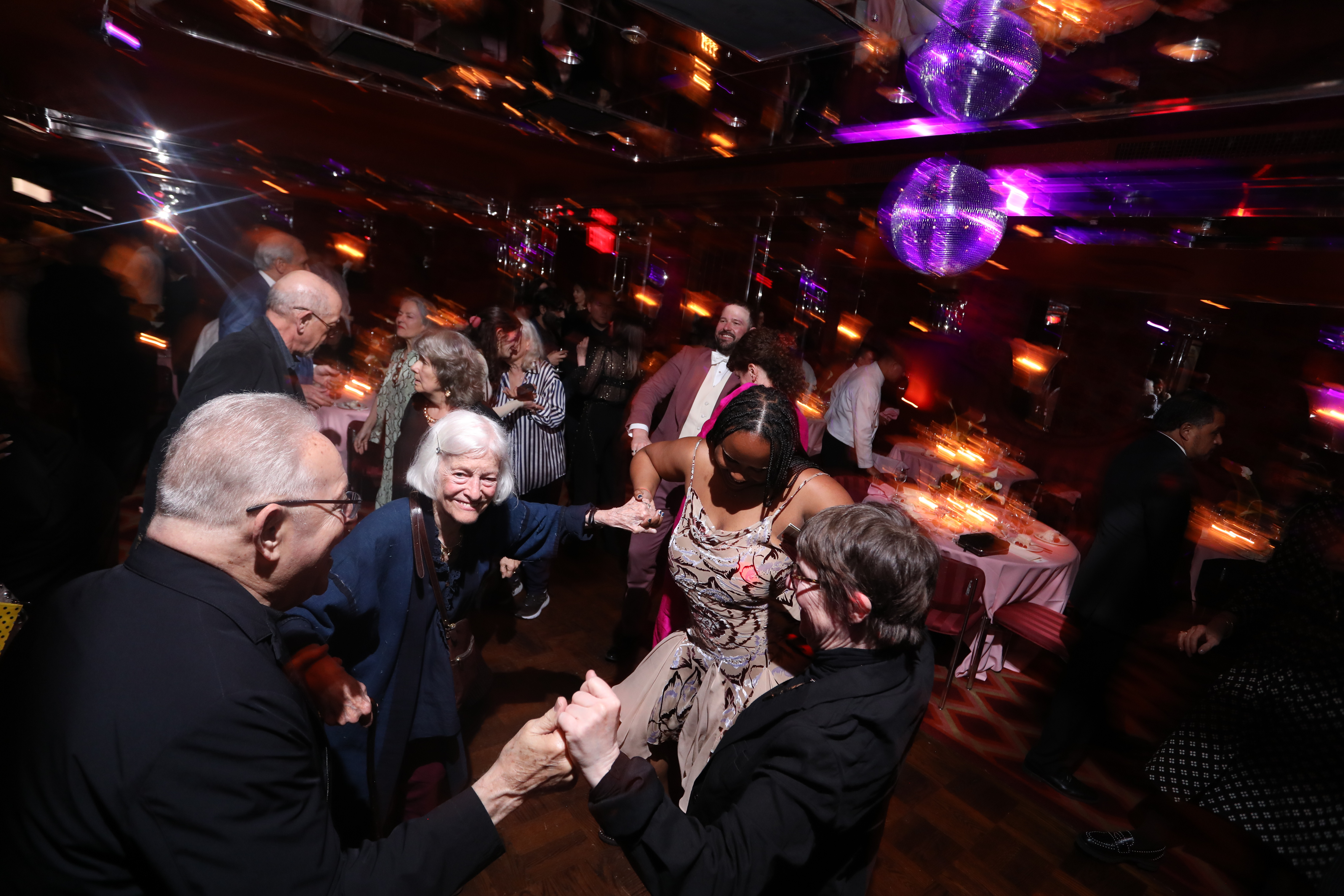 Color image of four individuals holding hands and dancing in a circle energetically. The background depicts a dimly lit venue with a glowing discoball and a variety of guests.