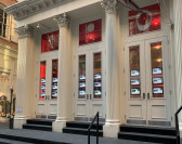 The facade of a cast-iron building with three double door entrances. Real estate listings are illuminated within the windows inside each door. In red tinted transoms above the doors are the letters N, R, O in eccentric white lettering.