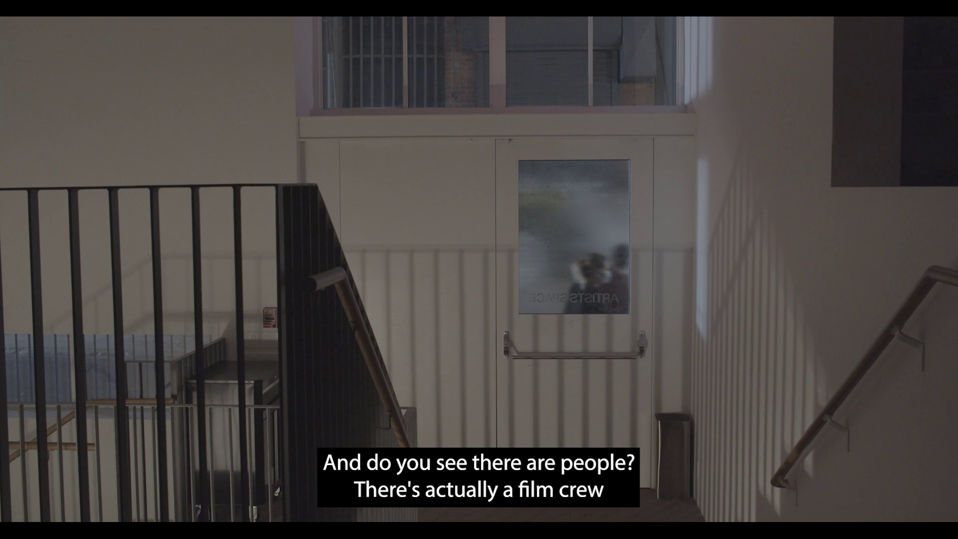 A set of stairs leads down to the entrance to Artists Space; the stair railings are cast in shadow on the wall. Through the door are two people and fake smoke. A caption appears over the image that reads: “And do you see there are people? There’s actually a film crew.”