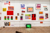An exhibition wall filled with colorful posters, prints and magazine covers, a tv monitor sits on the floor.
