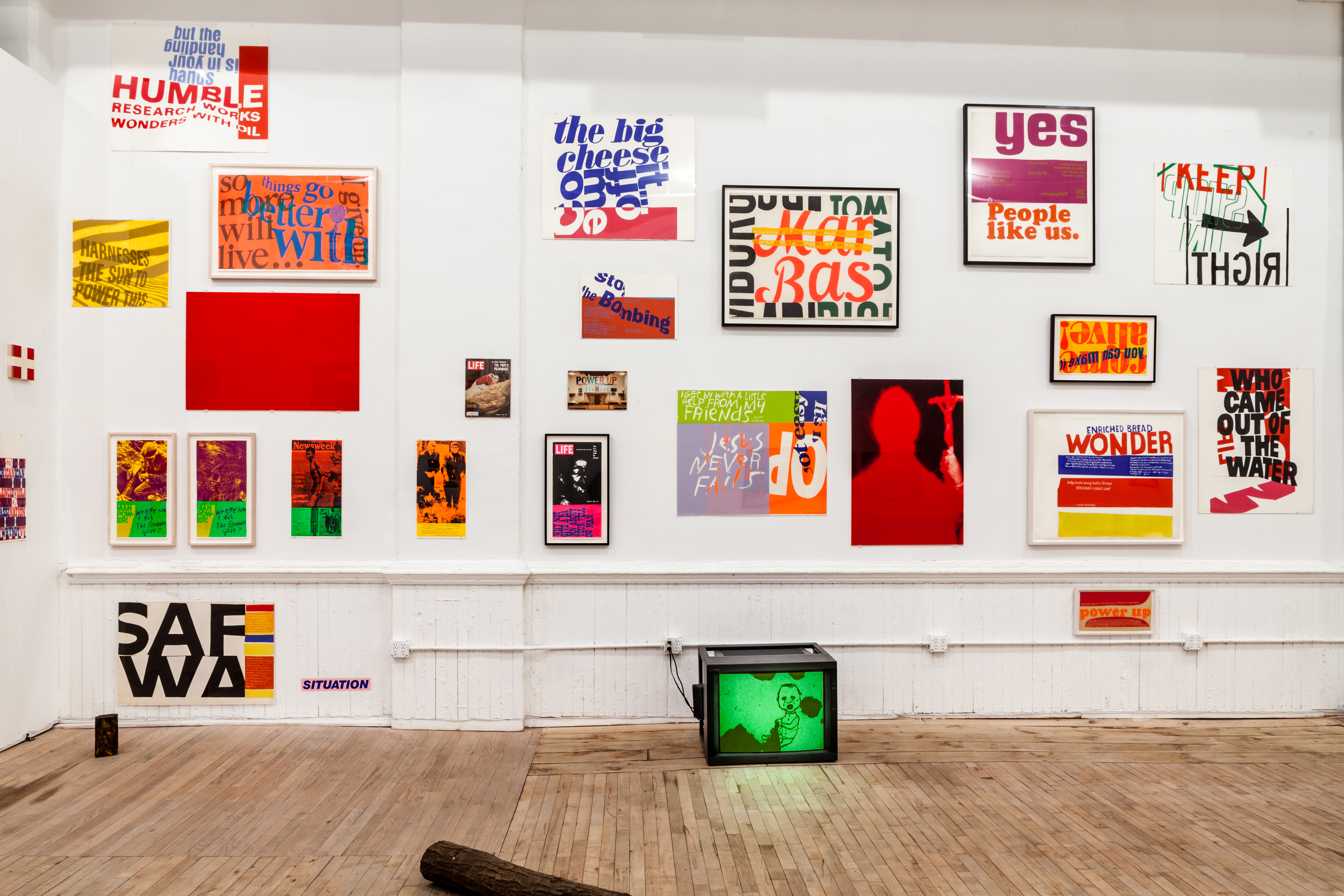 An exhibition wall filled with colorful posters, prints and magazine covers, a tv monitor sits on the floor.