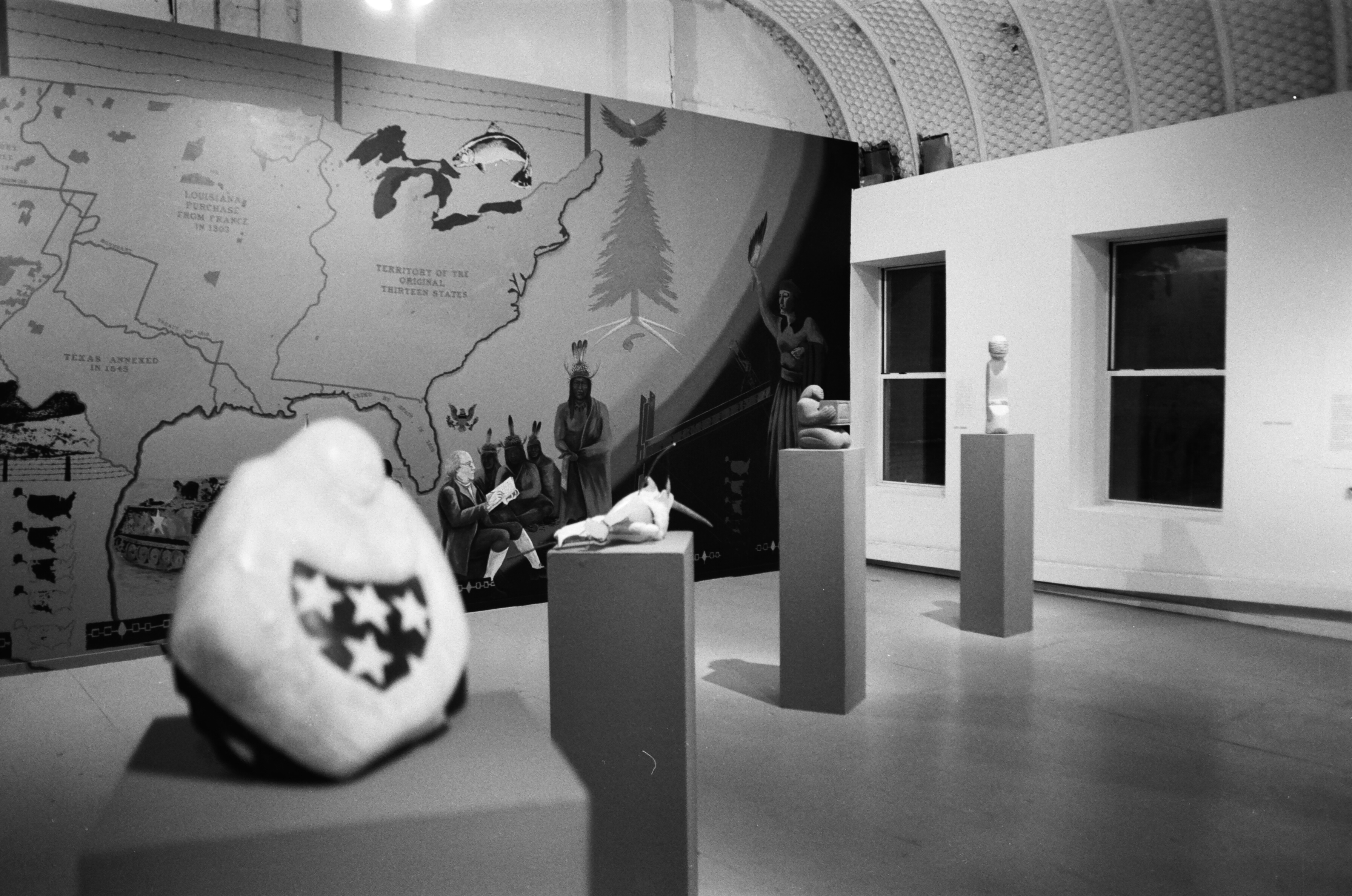 A black and white photo of an exhibition space, four sculptures on pedestals line the room diagonally with a mural painting behind them.