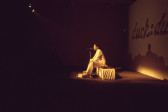 A color image of a person sitting on a pedestal, speaking into a standing microphone.