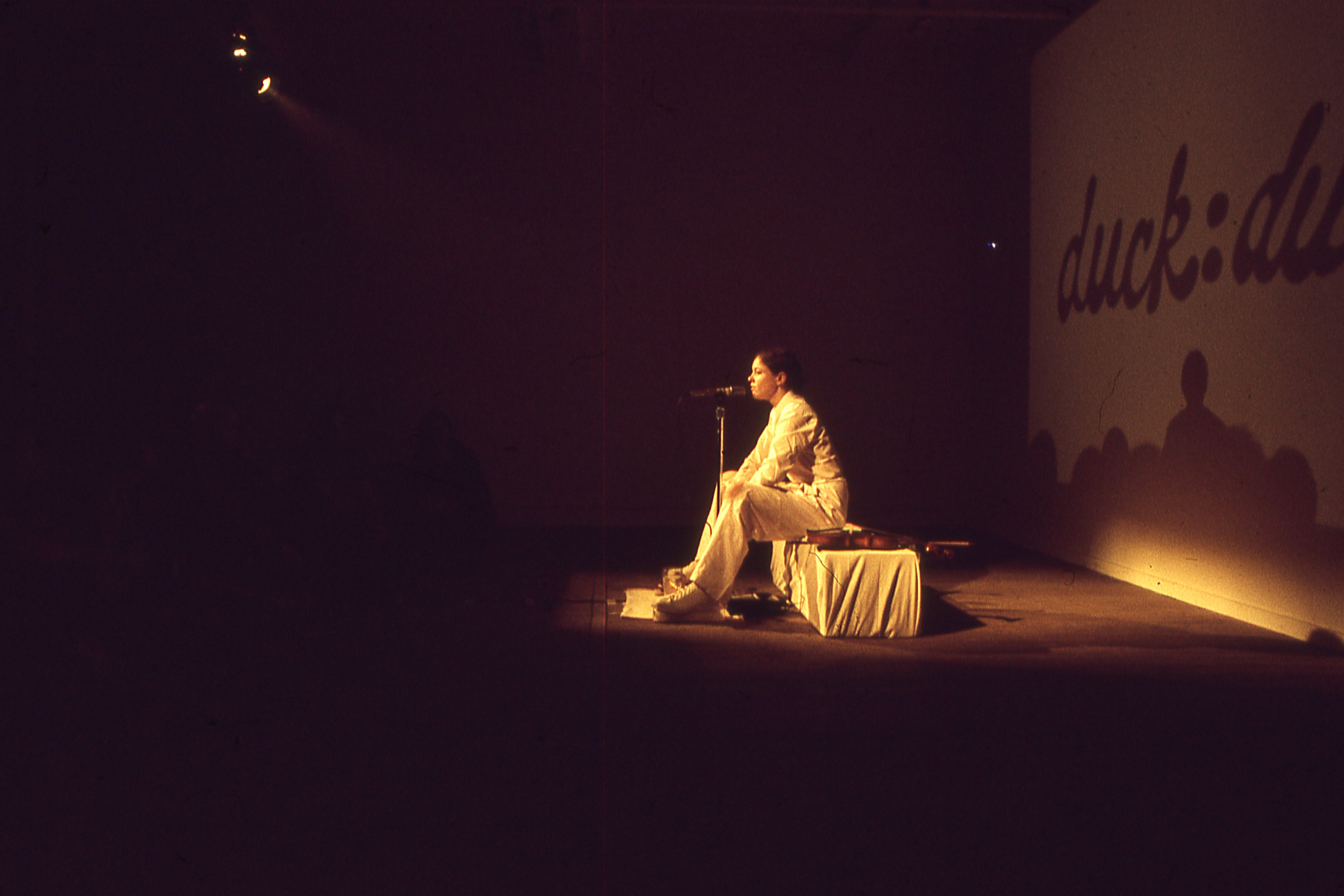 A color image of a person sitting on a pedestal, speaking into a standing microphone.