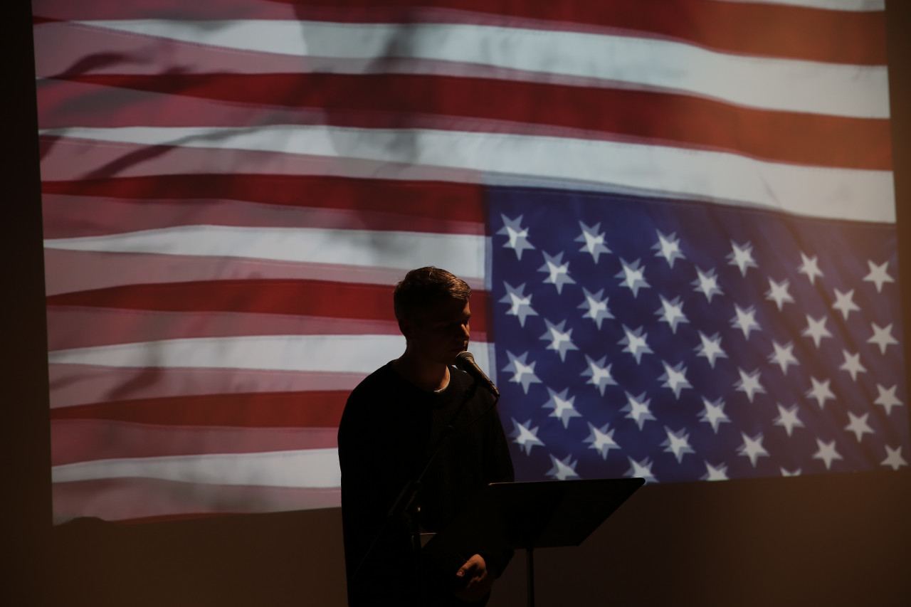 A figure stands in front of a music stand speaking into a microphone. They are backlit by an image of a rippling upside down American flag.