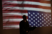 A figure stands in front of a music stand speaking into a microphone. They are backlit by an image of a rippling upside down American flag.