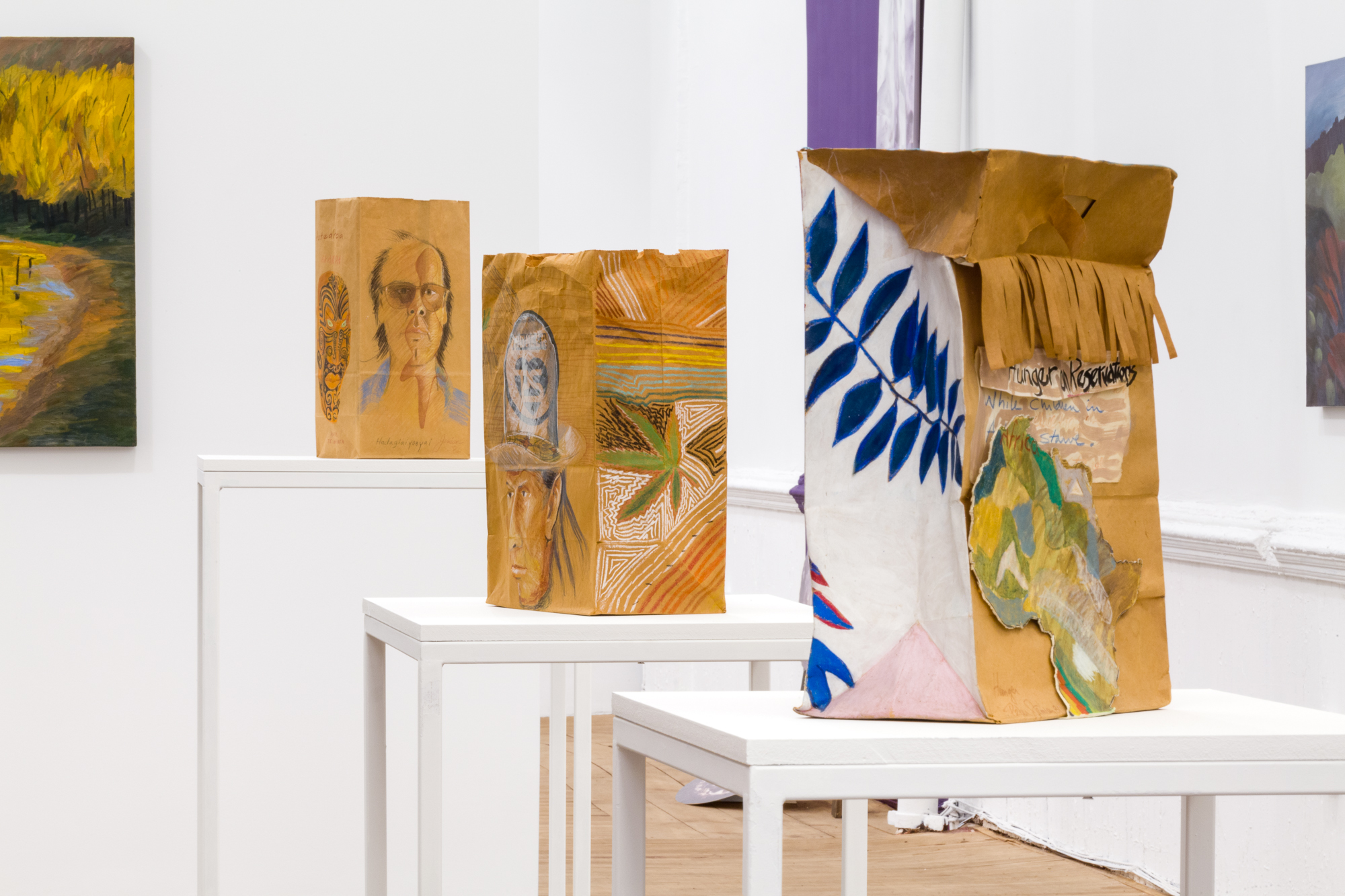 Three paper bags featuring collaged and drawn embellishments sit in a row on three pedastals of varying heights in ascending order.