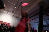 A person dressed in all pink extends a pink heart-shaped canvas over her head in front of a rapt audience. There are light spots reflected at even intervals patterning the room.