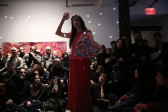 A person dressed in all pink holds a colorful heart-shaped canvas in her right hand while extending her left arm in the air. She is surrounded by rapt audience, who carefully track her movements.