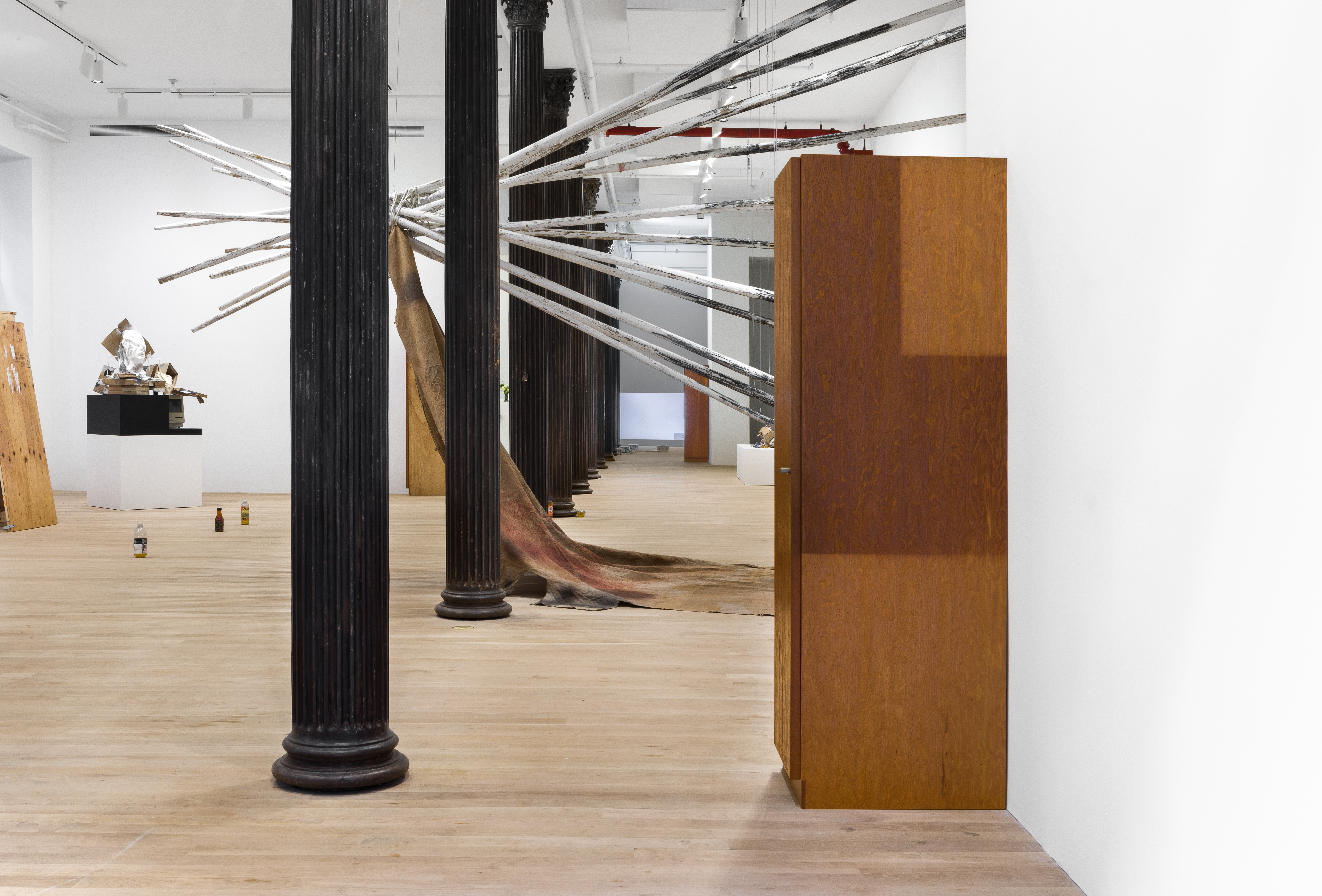 An installation view of several works in a gallery space. Twelve long wooden poles are mounted on a white gallery wall in a circular formation, projecting out from the wall and intersecting a line of black columns, forming a teepee shape. A large piece of brown linen stained with various black, white, and rust-colors hangs from the poles. Part of the linen drapes on the ground below the teepee structure. A wooden wardrobe stands against a wall in front of the structure. Several bottles filled with urine stand in a line behind the structure, in front of an abstract sculpture in the corner of the room. The sculpture contains a humanoid head, a TV monitor, and haphazard pieces of wooden and cardboard.