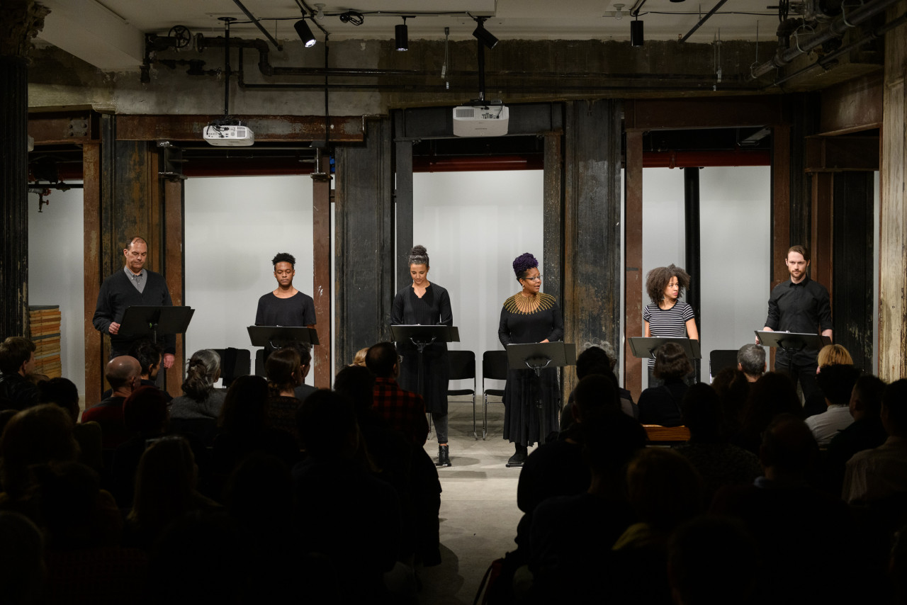A head-on view of six figures, each standing before a music stand in front of a seated audience. They are arranged side by side in a horizontal line, illuminated by overhead lights.