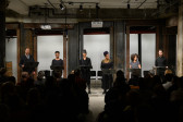 A head-on view of six figures, each standing before a music stand in front of a seated audience. They are arranged side by side in a horizontal line, illuminated by overhead lights.