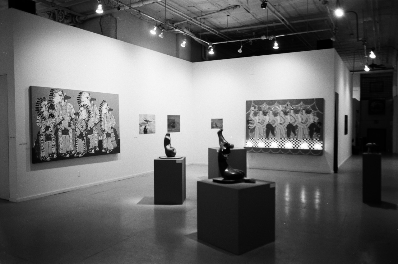 A black and white image of an exhibtion space, sculptures sit on three pedestals in front a wall of paintings.