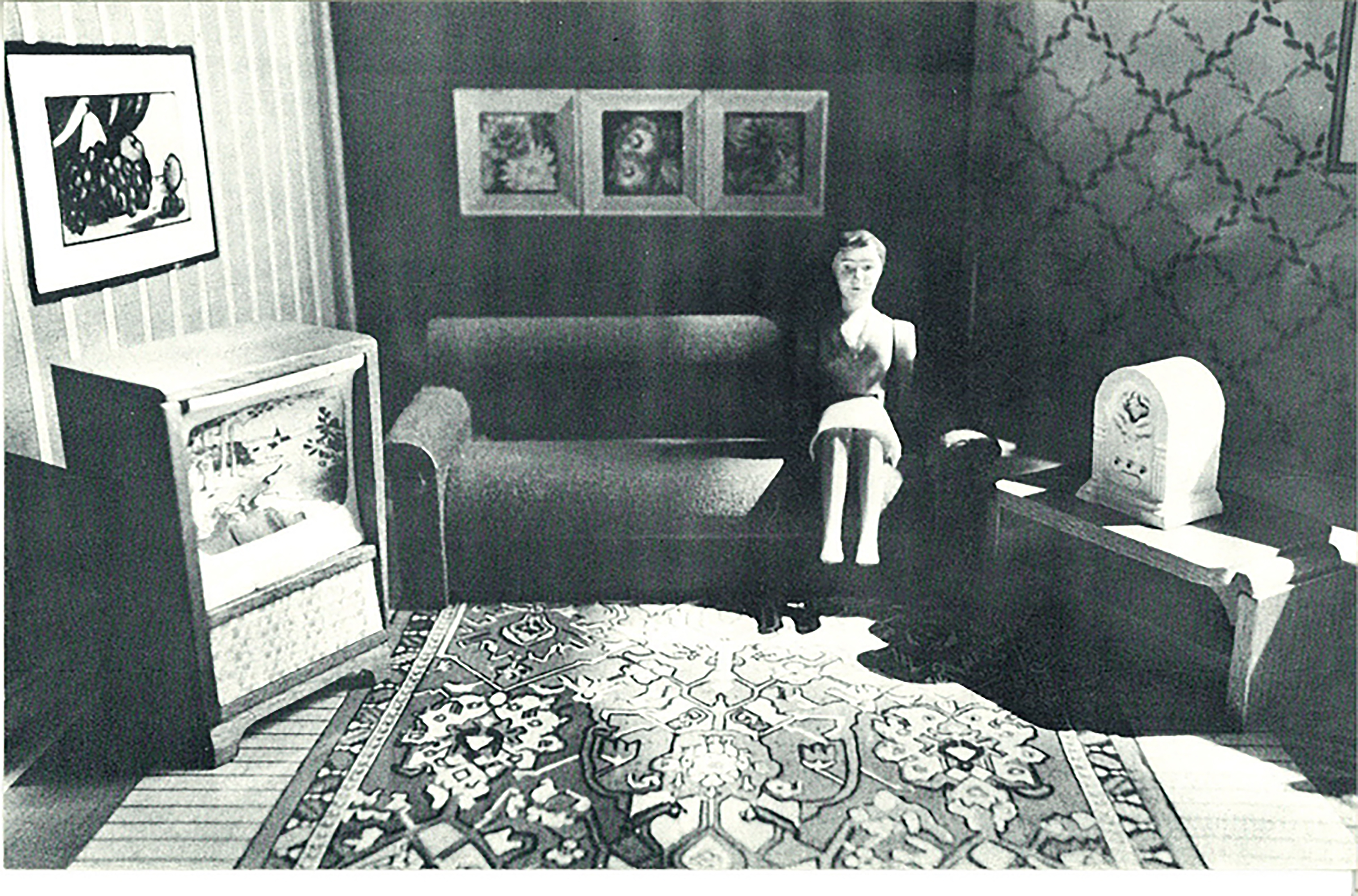 A black and white image of a dollhouse living room, a doll is sitting in the right corner, on a couch, next to a side table with radio on it.