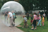 Two men walking inside a large inflatable sphere being followed by a crowd of people, some who are filming them, outside in a grassy and paved area.