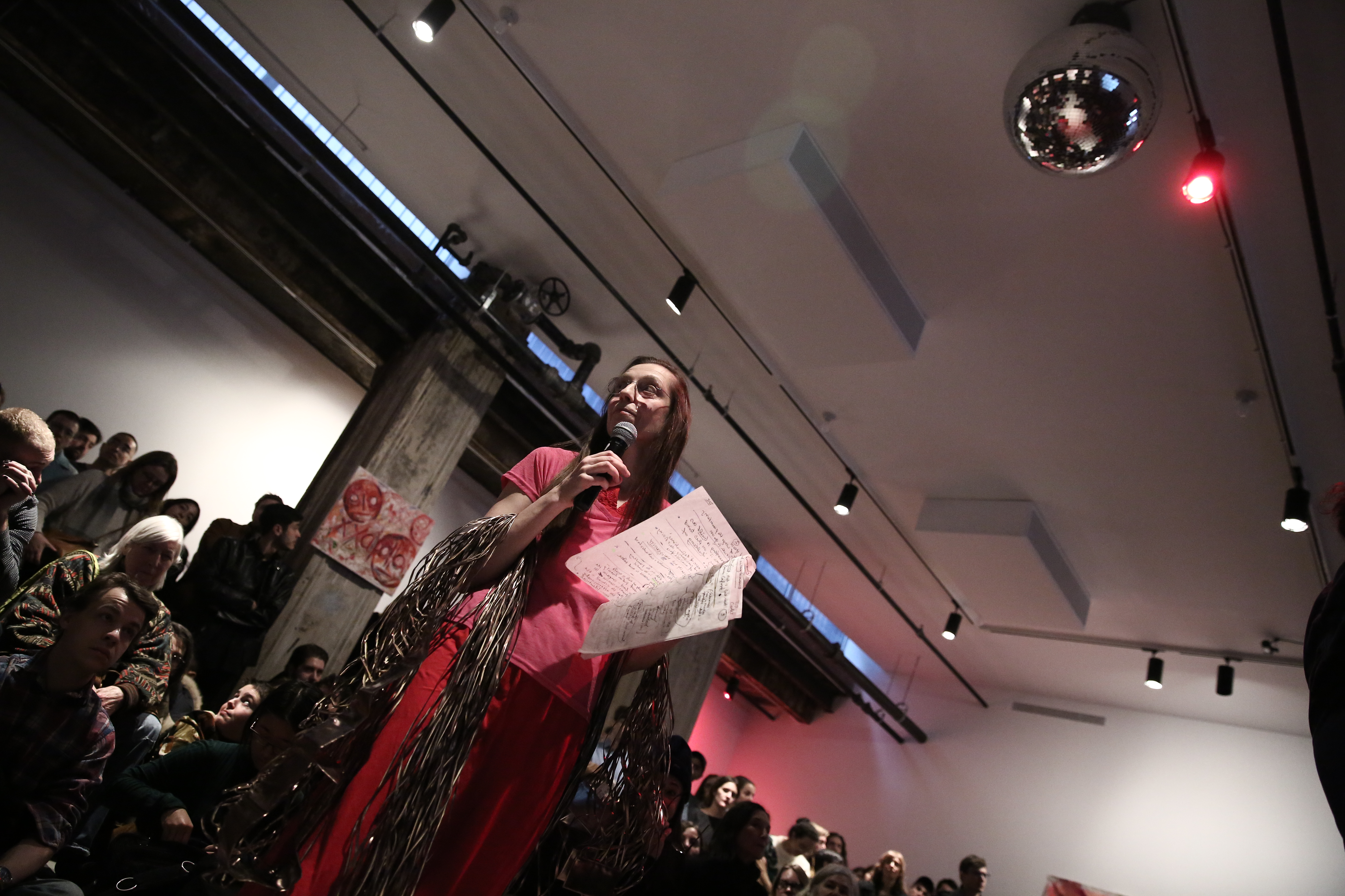A person dressed in all pink, holding several sheets of paper, speaks into a microphone, her arms are draped with masses of cords. A crowd of audience members surrounds her, some seated and some standing.