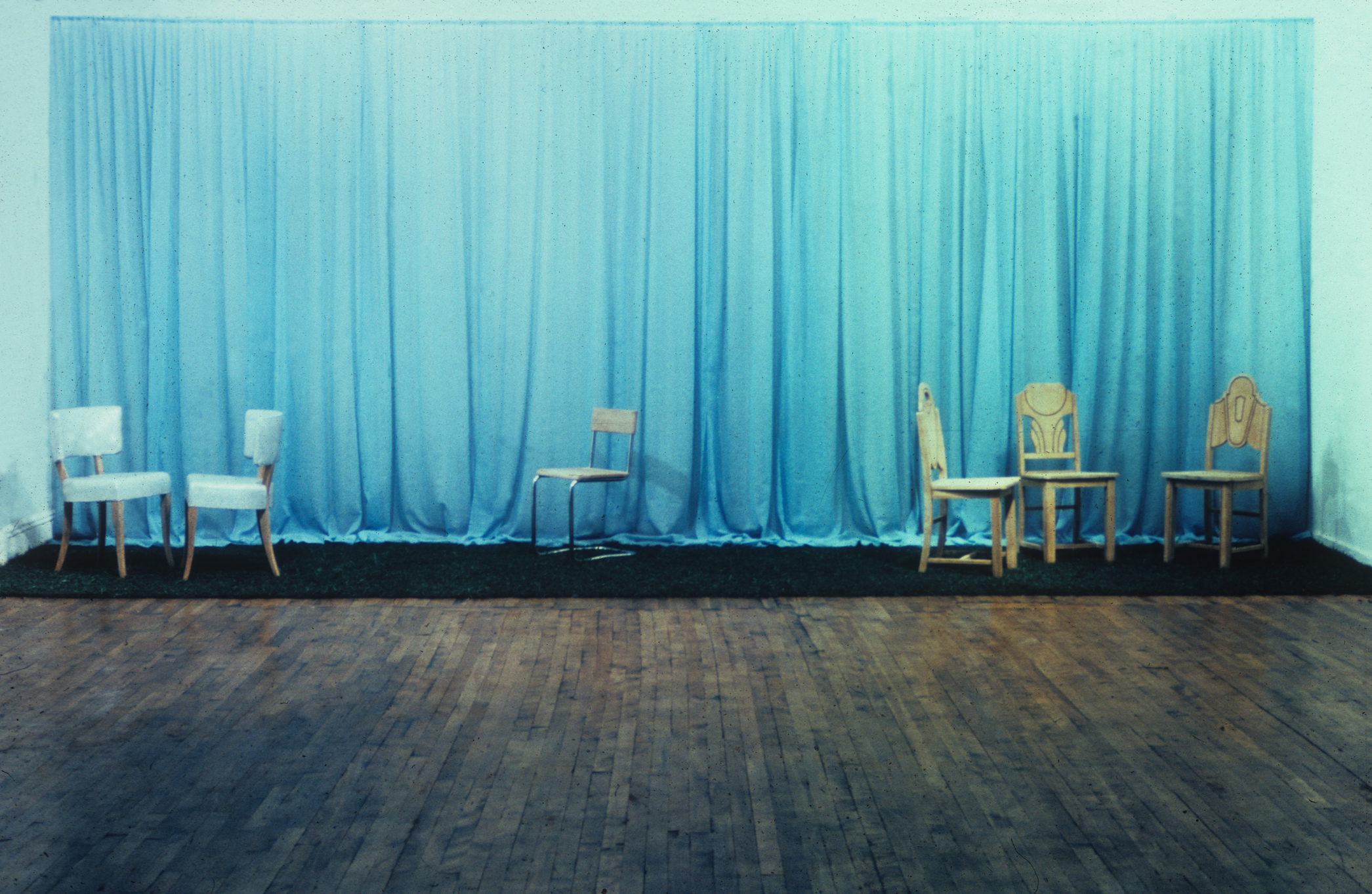 A stage set with a blue curtain, in front of which sit 6 chairs facing different directions.