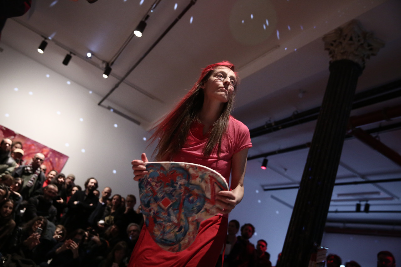 A person dressed in all pink grips a heart-shaped canvas in front of her, while her eyes look past the camera. She is surrounded by rapt audience members behind her. There are light spots reflected at even intervals patterning the room.