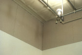 In the corner of a room, close to the ceiling, is an industrial light fixture. The lightbulb is on and faces up towards the ceiling.