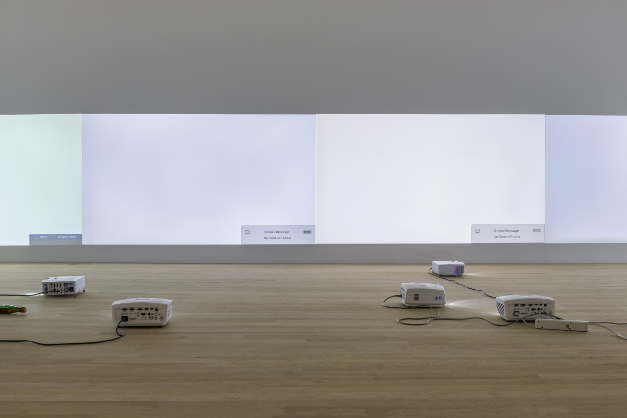 Projectors placed across a wood floor project variegated hues of white light onto a wall.