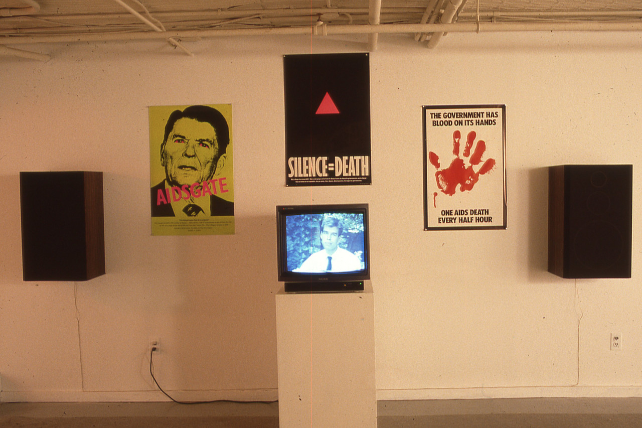 An exhibition wall with multiple posters hanging in between two large speakers and a tv on a pedestal in the center.