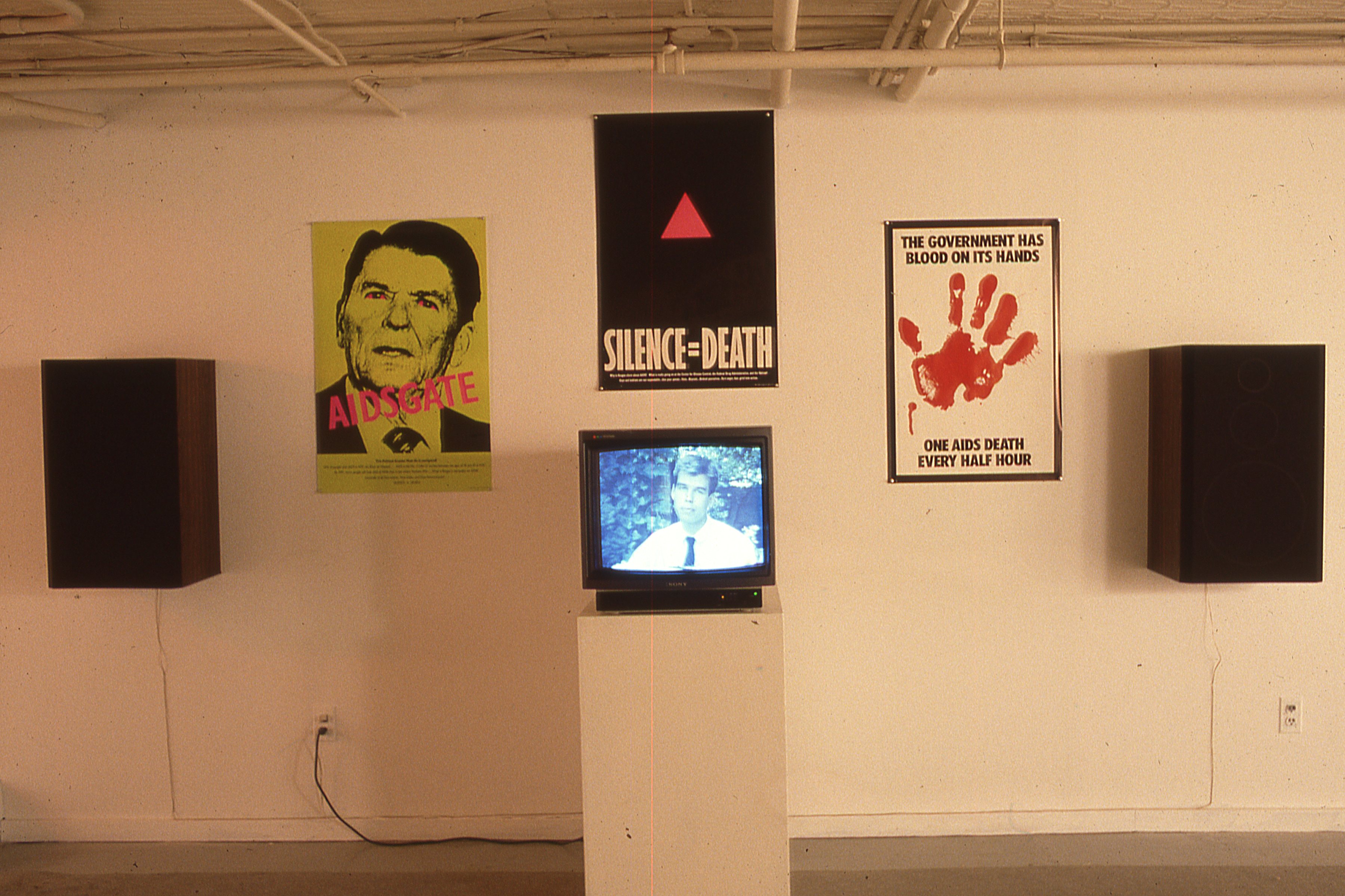 An exhibition wall with multiple posters hanging in between two large speakers and a tv on a pedestal in the center.